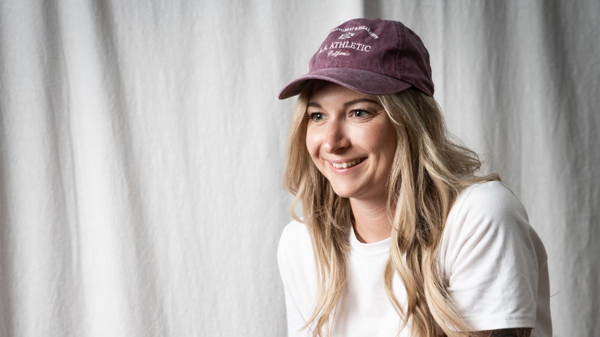 Kerstin Buber lacht in die Kamera, mit Beere-farbener Cap und weißem Shirt vor einem hellen Stoffhintergrund